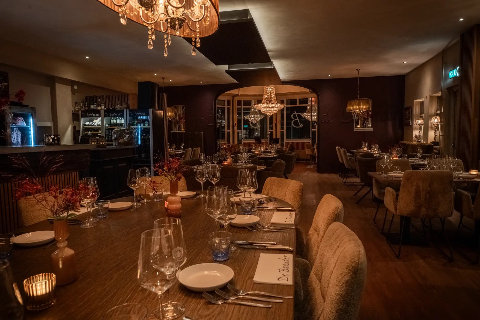Lange gedekte tafel met wijnglazen en kaarslicht, kroonluchter boven en bar op de achtergrond