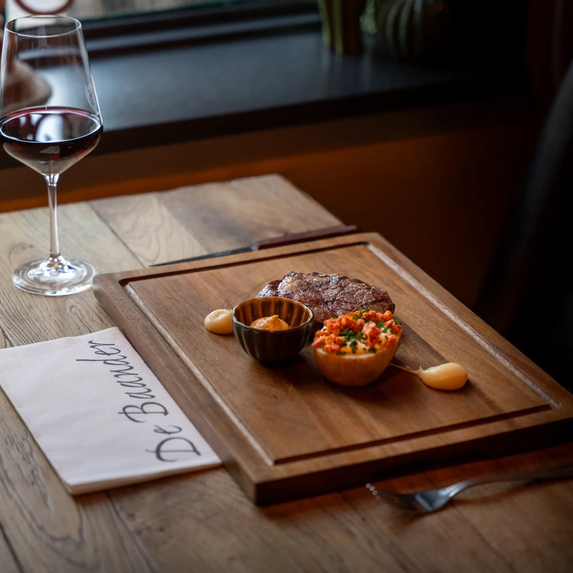 Gegrilde steak op houten plank met sausjes, naast een glas rode wijn en servet van De Baander