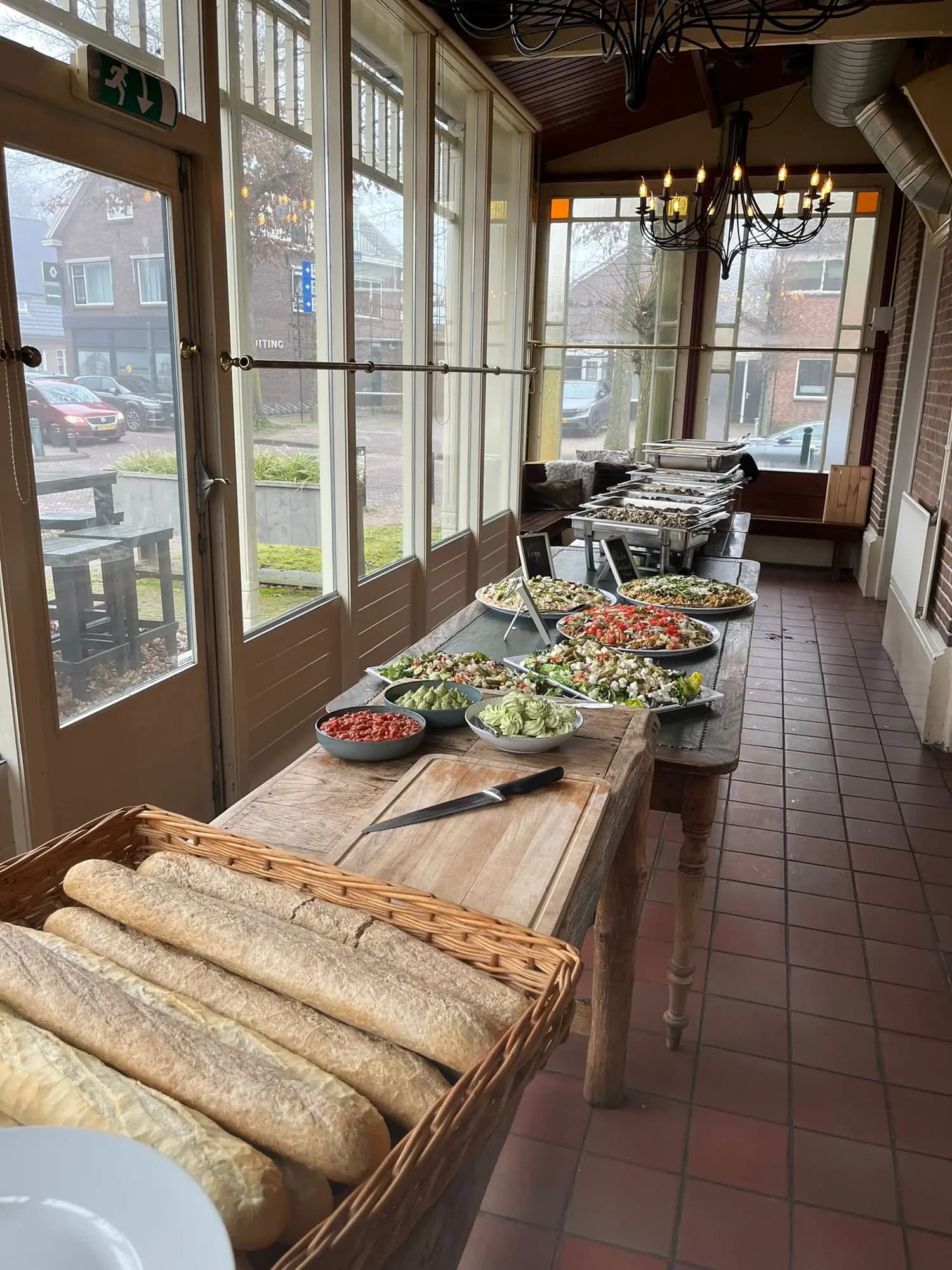 Buffettafel met verse stokbroden, salades en schalen langs de ramen van een lichte serre