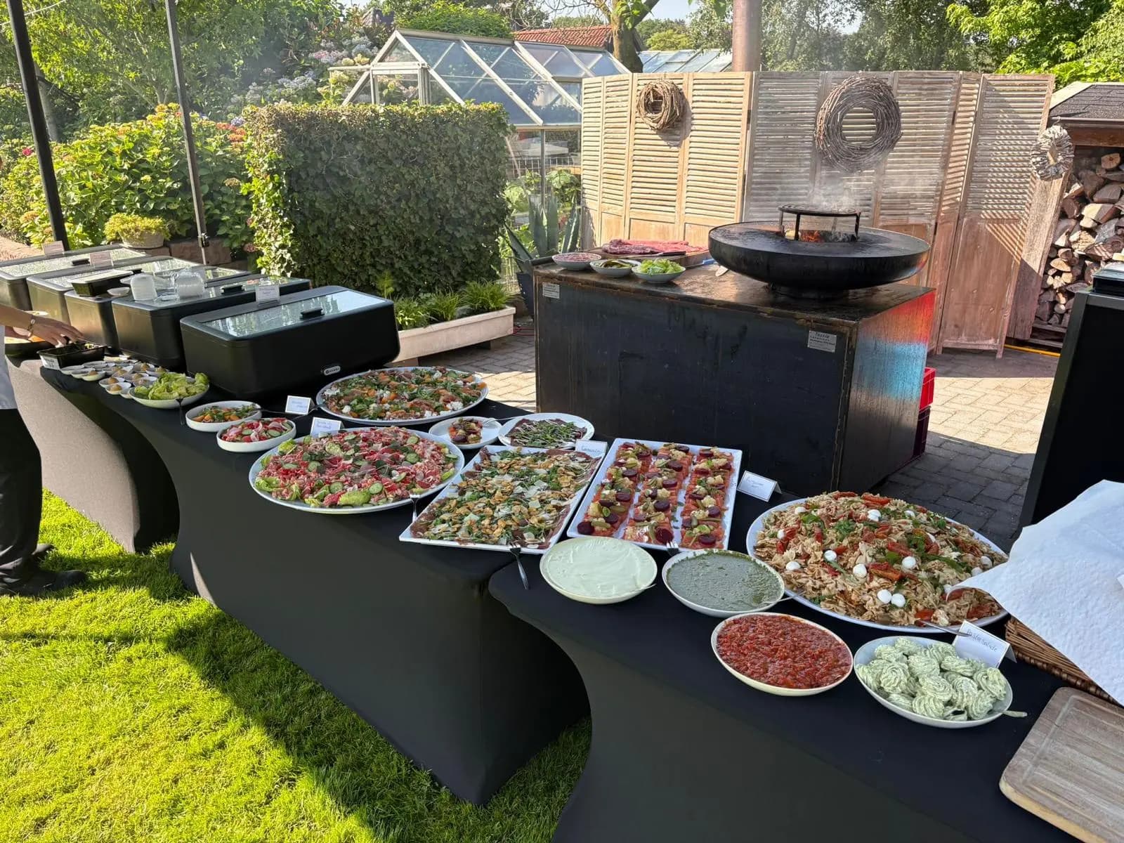 Buitenbuffet met salades en plateaus op zwarte tafels, met vuurschaal in een tuin