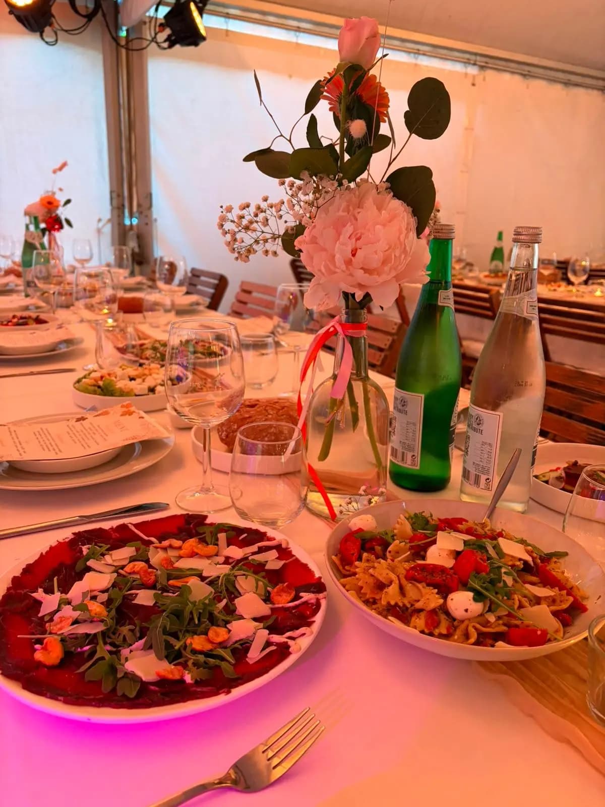 Gedekte feesttafel met carpaccio, pastasalade, bloemen en water in een feesttent
