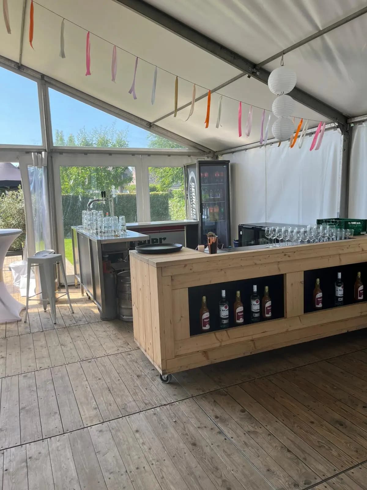 Houten mobiele bar met tapinstallatie, glazen en koeling in een feesttent met slingers