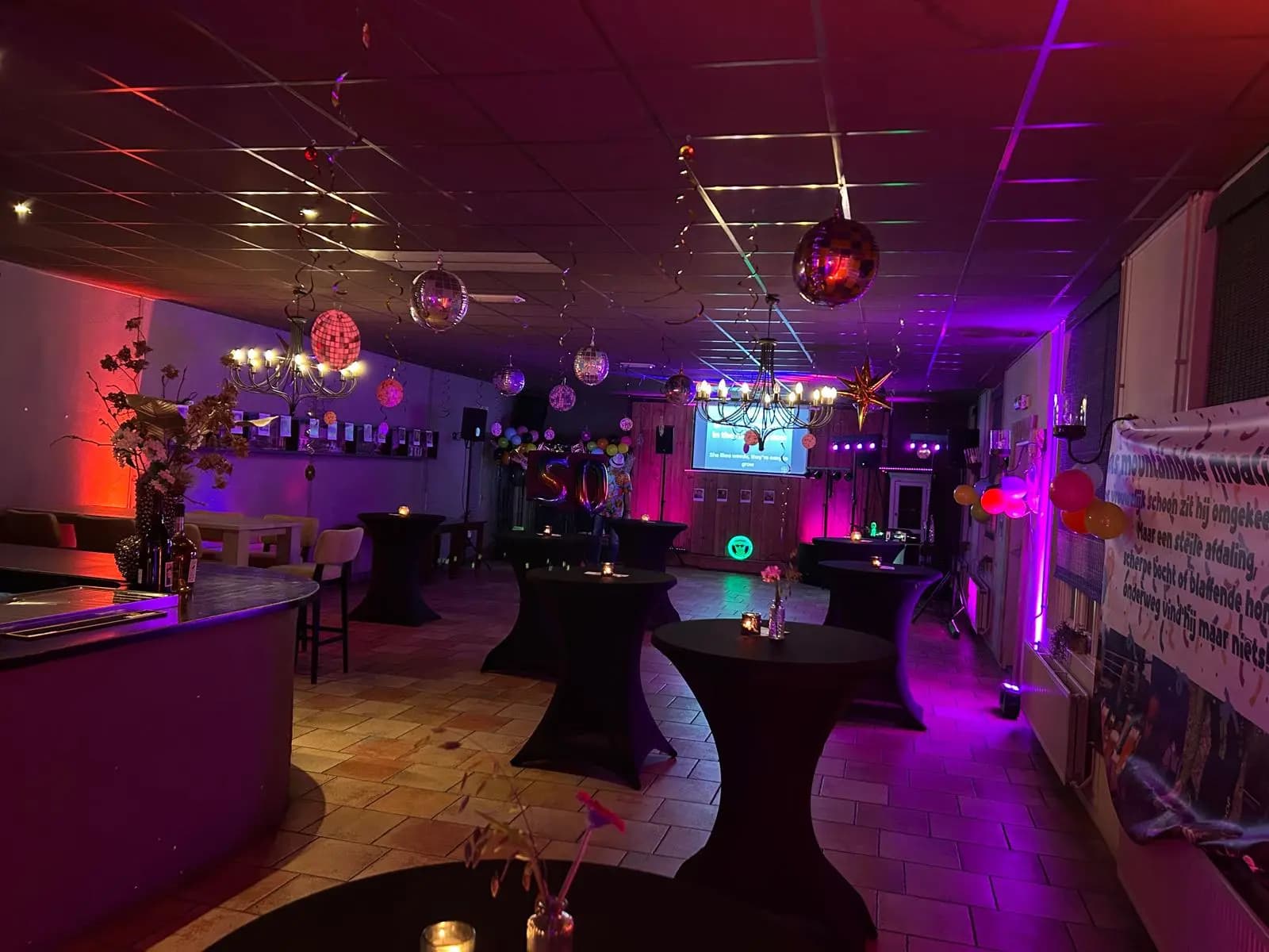 Feestzaal met discoballen, statafels en paarse sfeerverlichting, ingericht voor een vijftigste verjaardag