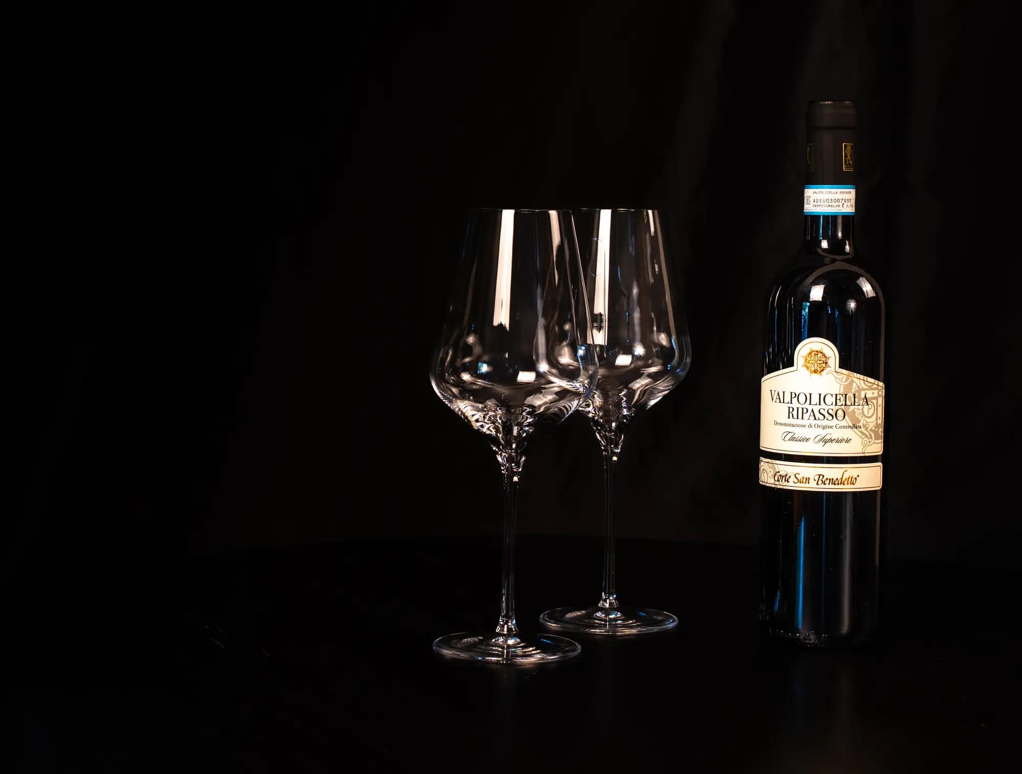 Fles Valpolicella Ripasso Classico Superiore naast twee lege wijnglazen op een donkere tafel