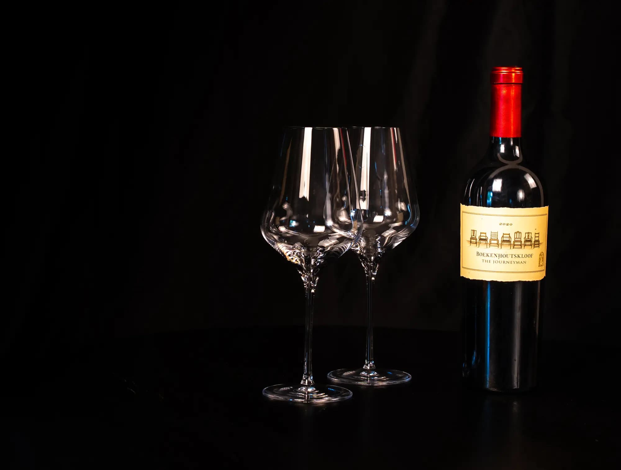 Fles Boekenhoutskloof The Journeyman naast twee lege wijnglazen op een donkere tafel