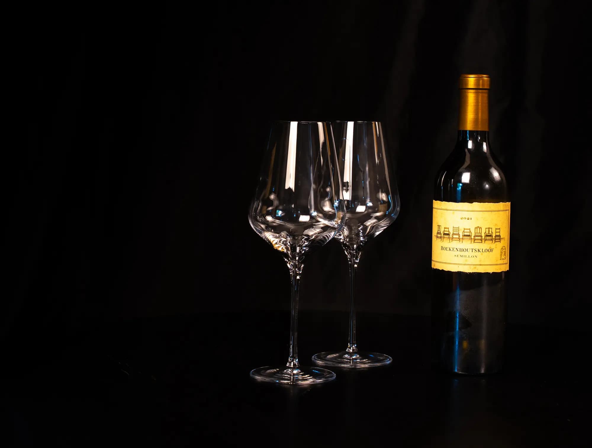 Fles Boekenhoutskloof Semillon naast twee lege wijnglazen op een donkere tafel
