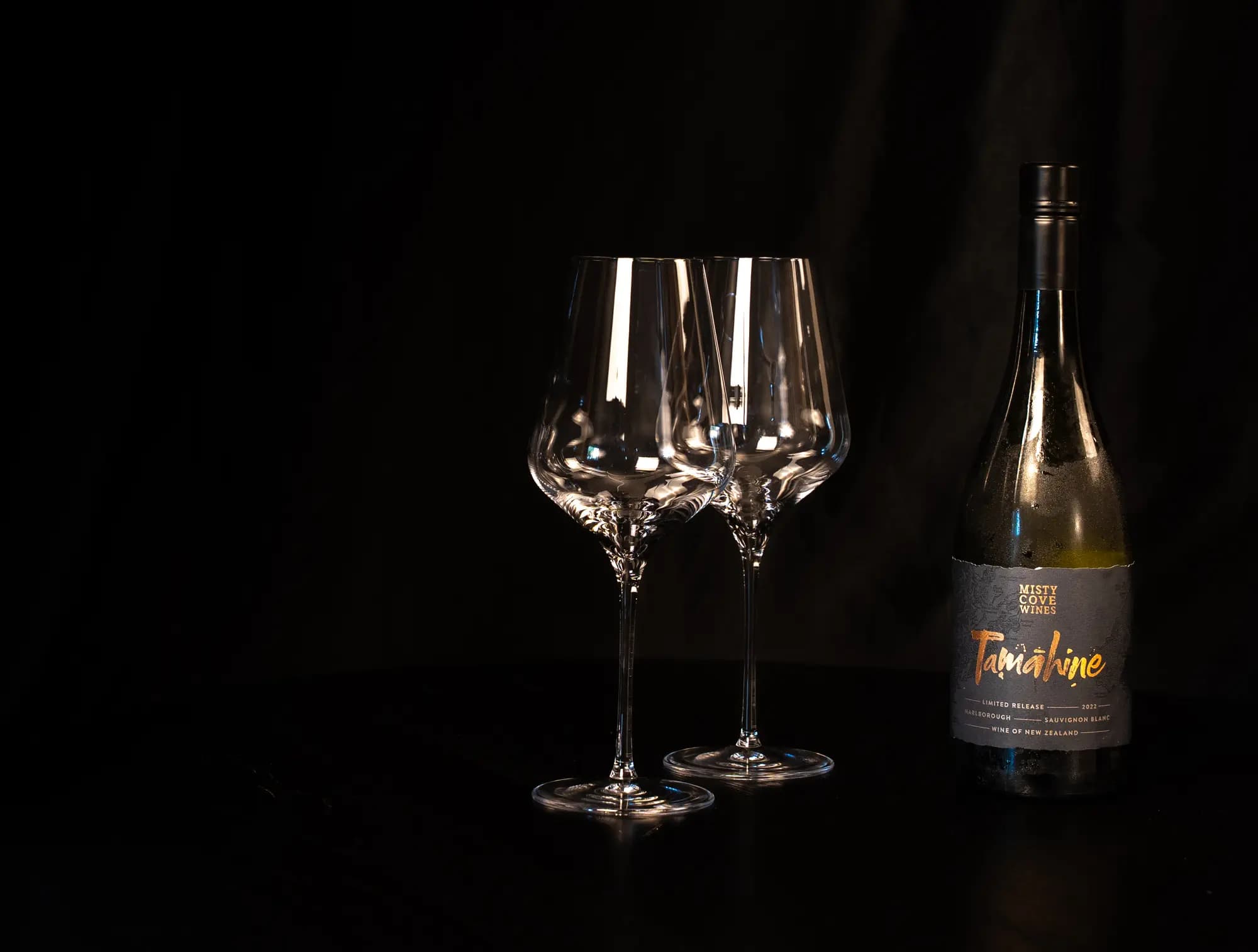 Fles Misty Cove Tamahine Sauvignon Blanc naast twee lege wijnglazen op een donkere tafel