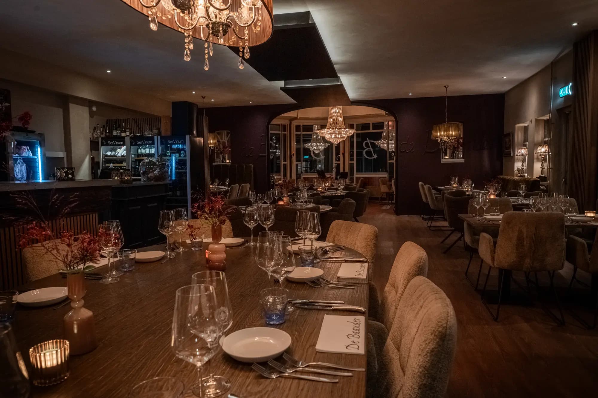 Gedekte tafels en kroonluchters in het avondlijk verlichte restaurant de Baander, met de bar op de achtergrond