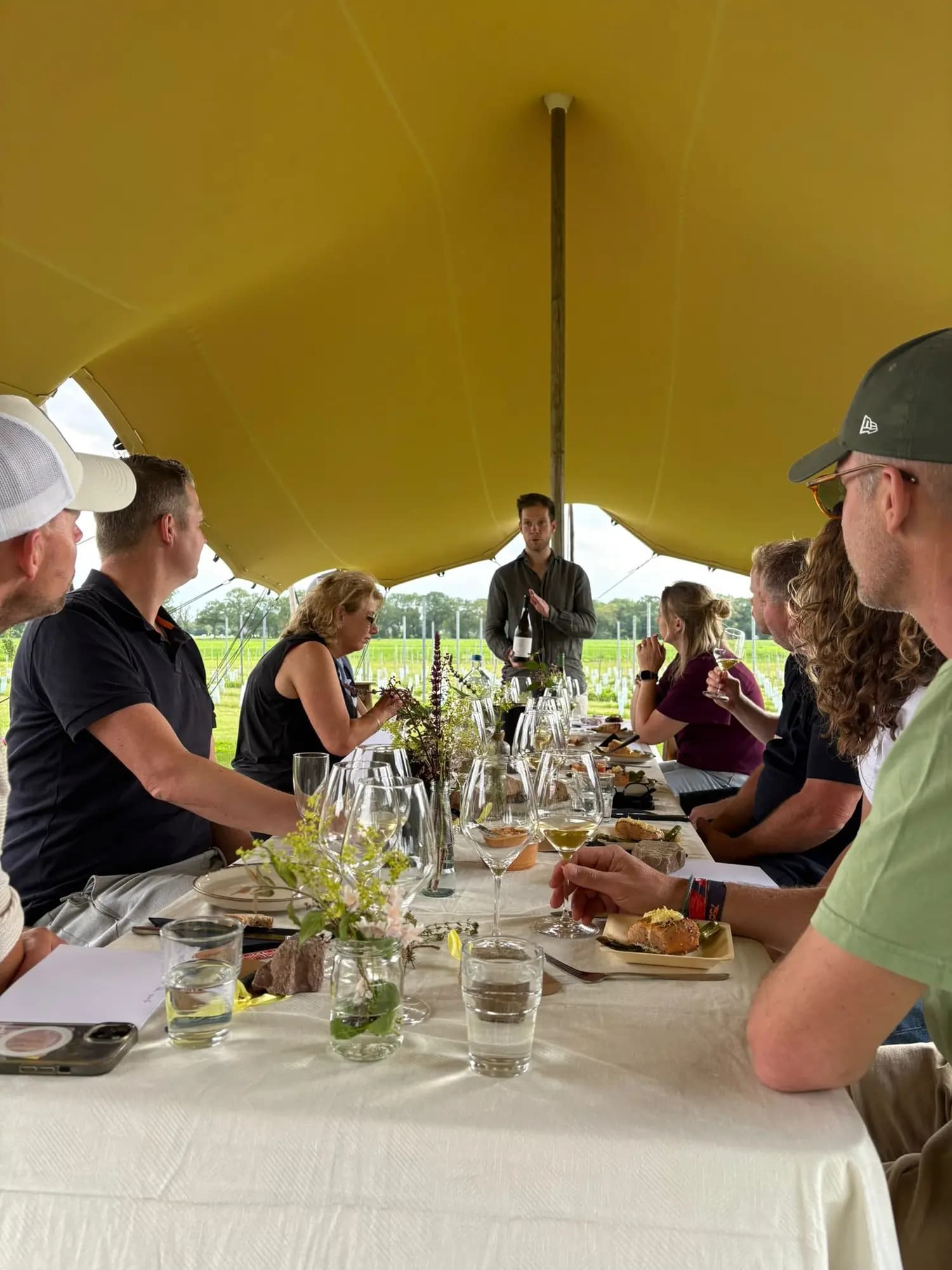 Gezelschap aan een lange tafel tijdens een wijnproeverij onder een gele tent bij de wijngaard